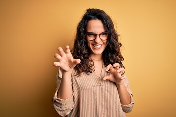 Beautiful woman with curly hair wearing striped shirt and glasses over yellow background smiling...