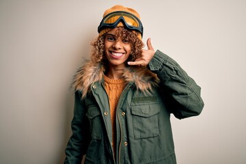 Young african american skier woman with curly hair wearing snow sportswear and ski goggles smiling doing phone gesture with hand and fingers like talking on the telephone. Communicating concepts.
