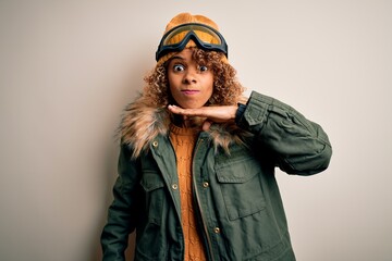 Young african american skier woman with curly hair wearing snow sportswear and ski goggles cutting throat with hand as knife, threaten aggression with furious violence