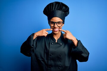 Young african american chef woman wearing cooker uniform and hat over blue background smiling cheerful showing and pointing with fingers teeth and mouth. Dental health concept.