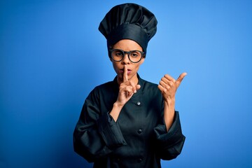 Young african american chef woman wearing cooker uniform and hat over blue background asking to be quiet with finger on lips pointing with hand to the side. Silence and secret concept.