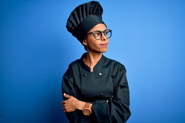 Young african american chef woman wearing cooker uniform and hat over blue background looking to the side with arms crossed convinced and confident