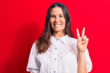 Young beautiful brunette woman wearing casual shirt standing over isolated red background smiling with happy face winking at the camera doing victory sign. Number two.