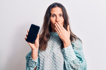 Young beautiful brunette woman holding smartphone showing screen over white background covering...