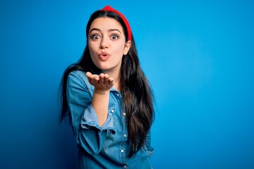 Young brunette woman wearing casual denim shirt over blue isolated background looking at the camera blowing a kiss with hand on air being lovely and sexy. Love expression.