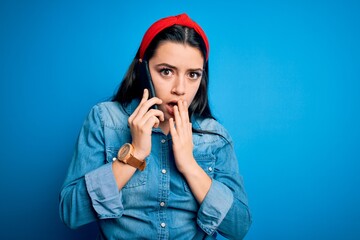 Young brunette woman talking on smartphone over blue isolated background cover mouth with hand...