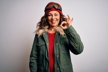 Middle age skier woman wearing snow sportswear and ski goggles over white background smiling positive doing ok sign with hand and fingers. Successful expression.