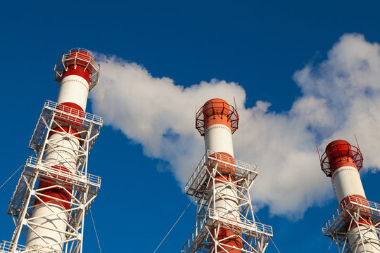 Three striped red and white factory chimneys from which white smoke comes out, stand against a blue sky.