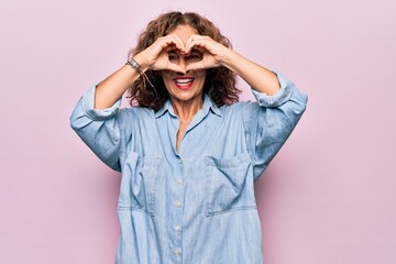 Middle age beautiful woman wearing casual denim shirt standing over pink background Doing heart...