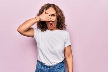 Middle age beautiful woman wearing casual t-shirt standing over isolated pink background peeking in shock covering face and eyes with hand, looking through fingers afraid