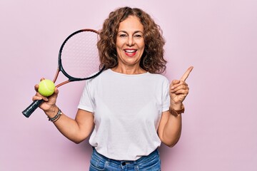 Middle age beautiful sportswoman playing tennis holding racket and ball over white background smiling happy pointing with hand and finger to the side