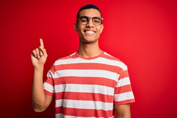 Young handsome african american man wearing casual striped t-shirt and glasses with a big smile on face, pointing with hand finger to the side looking at the camera.