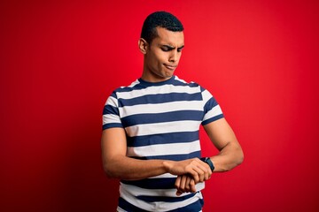 Handsome african american man wearing casual striped t-shirt standing over red background Checking the time on wrist watch, relaxed and confident