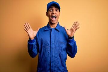 Young african american mechanic man wearing blue uniform and cap over yellow background celebrating crazy and amazed for success with arms raised and open eyes screaming excited. Winner concept