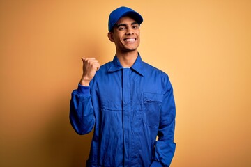 Young african american mechanic man wearing blue uniform and cap over yellow background smiling with happy face looking and pointing to the side with thumb up.