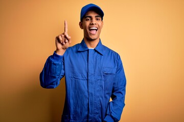 Young african american mechanic man wearing blue uniform and cap over yellow background showing and...