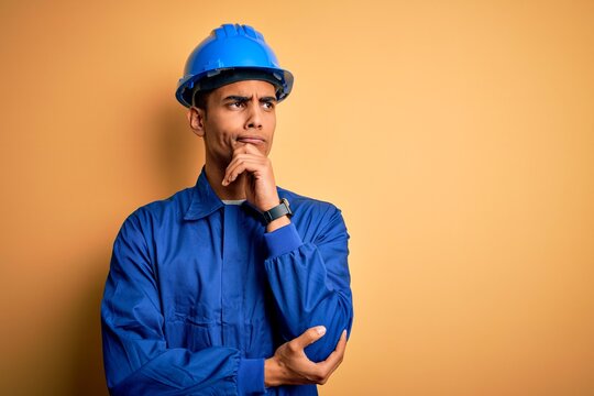 Young handsome african american worker man wearing blue uniform and security helmet with hand on chin thinking about question, pensive expression. Smiling with thoughtful face. Doubt concept.
