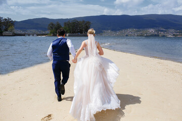 the bride and groom are running along the sandy beach, on the wedding day the newlyweds are having fun, a man holds his womans hand and runs near the ocean