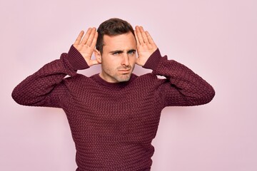 Young handsome man with blue eyes wearing casual sweater standing over pink background Trying to hear both hands on ear gesture, curious for gossip. Hearing problem, deaf