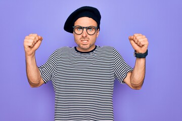 Handsome man with blue eyes wearing striped t-shirt and french beret over purple background angry...