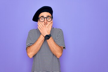 Handsome man with blue eyes wearing striped t-shirt and french beret over purple background shocked covering mouth with hands for mistake. Secret concept.