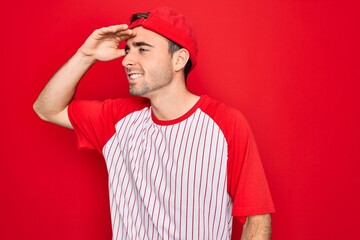 Young handsome sporty man with blue eyes wearing striped baseball t-shirt and cap very happy and smiling looking far away with hand over head. Searching concept.