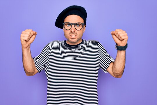 Handsome man with blue eyes wearing striped t-shirt and french beret over purple background angry and mad raising fists frustrated and furious while shouting with anger. Rage and aggressive concept.