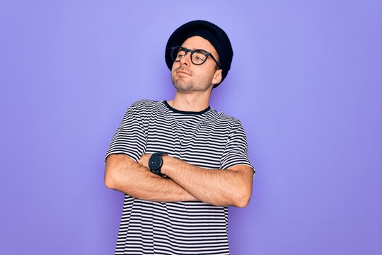 Handsome man with blue eyes wearing striped t-shirt and french beret over purple background looking to the side with arms crossed convinced and confident