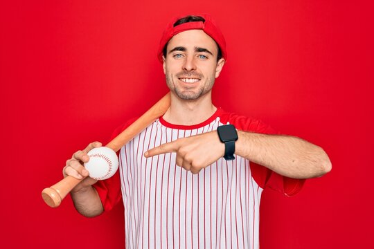 Young handsome sporty man with blue eyes playing baseball wearing cap holding bat and ball very happy pointing with hand and finger