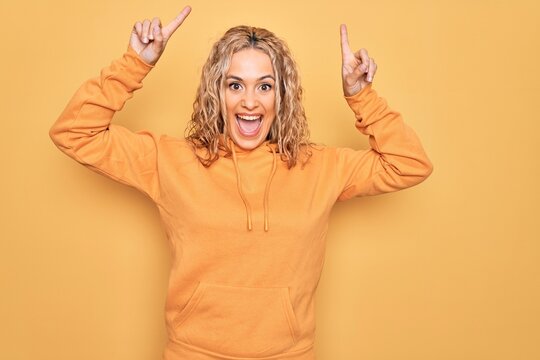 Young beautiful blonde sporty woman wearing casual sweatshirt over yellow background smiling amazed and surprised and pointing up with fingers and raised arms.
