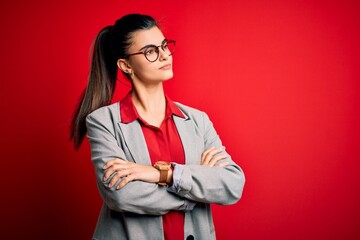 Young beautiful brunette businesswoman wearing jacket and glasses over red background looking to the side with arms crossed convinced and confident