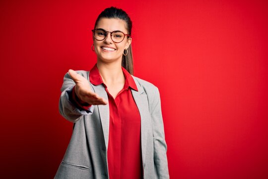 Young beautiful brunette businesswoman wearing jacket and glasses over red background smiling cheerful offering palm hand giving assistance and acceptance.
