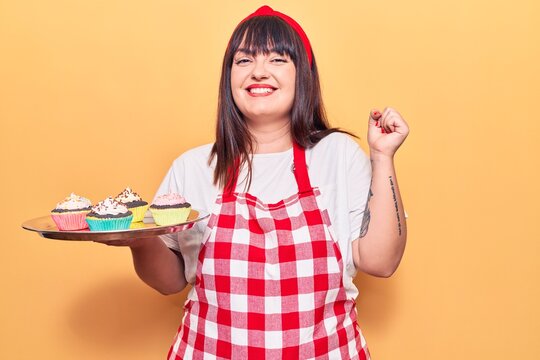 Young plus size woman wearing apron holding cupcake screaming proud, celebrating victory and success very excited with raised arm - Powered by Adobe