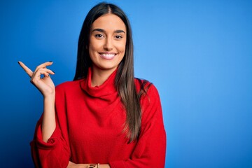 Young beautiful brunette woman wearing red casual sweater over isolated blue background with a big smile on face, pointing with hand and finger to the side looking at the camera.