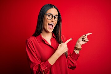 Naklejka premium Young beautiful brunette woman wearing casual shirt and glasses over red background smiling and looking at the camera pointing with two hands and fingers to the side.