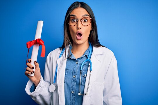 Young beautiful brunette doctor woman wearing glasses and coat holding diploma degree scared in shock with a surprise face, afraid and excited with fear expression