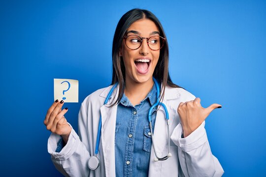 Young beautiful brunette doctor woman holding paper with question mark symbol message pointing and showing with thumb up to the side with happy face smiling