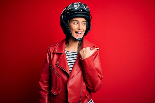 Young beautiful brunette motorcyclist woman wearing motorcycle helmet and red jacket smiling with happy face looking and pointing to the side with thumb up.