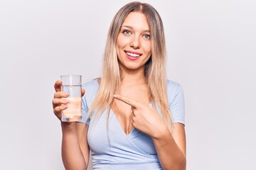 Young beautiful blonde girl drinking glass of water smiling happy pointing with hand and finger