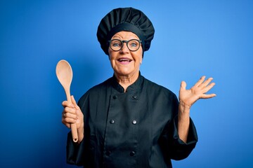 Naklejka premium Senior beautiful grey-haired chef woman wearing cooker uniform and hat holding wooden spoon very happy and excited, winner expression celebrating victory screaming with big smile and raised hands