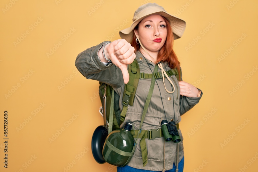 Poster Young redhead backpacker woman hiking wearing backpack and hat over yellow background looking unhappy and angry showing rejection and negative with thumbs down gesture. Bad expression.