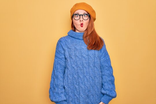 Young beautiful redhead woman wearing french beret and glasses over yellow background making fish face with lips, crazy and comical gesture. Funny expression.
