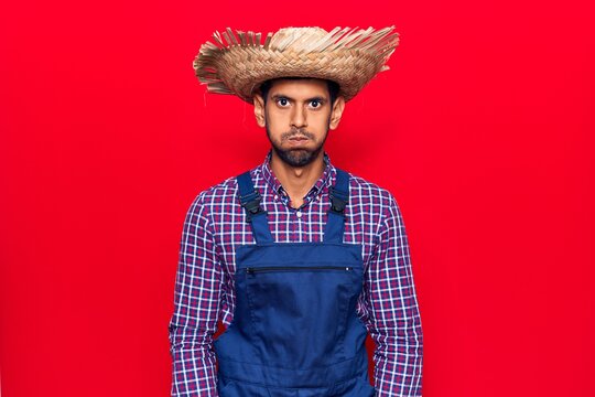 Young latin man wearing farmer hat and apron puffing cheeks with funny face. mouth inflated with air, crazy expression.