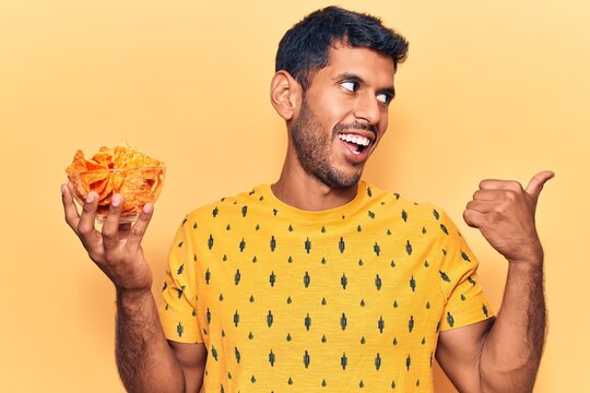 Young latin man holding nachos potato chips pointing thumb up to the side smiling happy with open mouth