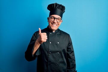 Middle age handsome grey-haired chef man wearing cooker uniform and hat doing happy thumbs up gesture with hand. Approving expression looking at the camera showing success.