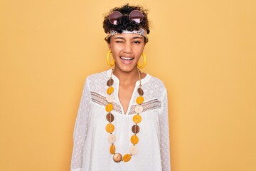 Young beautiful african american afro hippie woman wearing sunglasses and accessories winking...