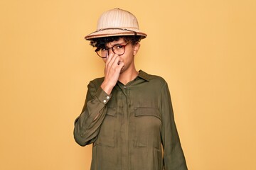 Young beautiful african american tourist woman wearing glasses and explorer hat smelling something...