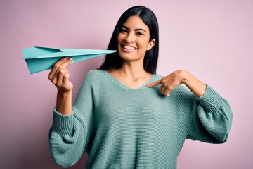 Young beautiful hispanic woman holding paper plane and communication and travel message with surprise face pointing finger to himself