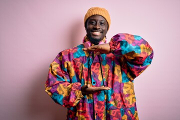 Obraz premium Young handsome african american man wearing colorful coat and cap over pink background gesturing with hands showing big and large size sign, measure symbol. Smiling looking at the camera. Measuring