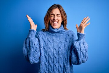 Young beautiful brunette woman wearing casual turtleneck sweater over blue background celebrating mad and crazy for success with arms raised and closed eyes screaming excited. Winner concept